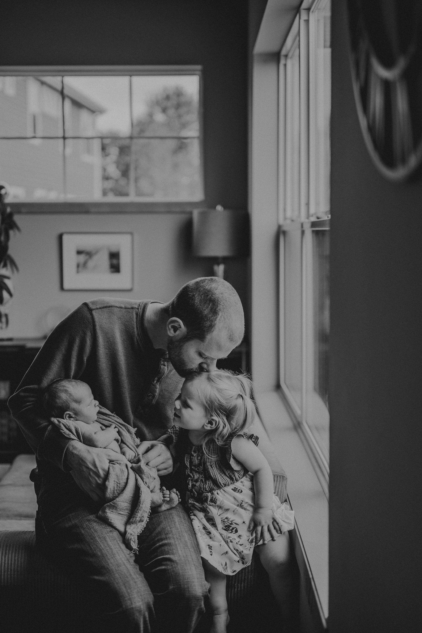 Black and white portrait of family bonding moment near window with natural lighting.