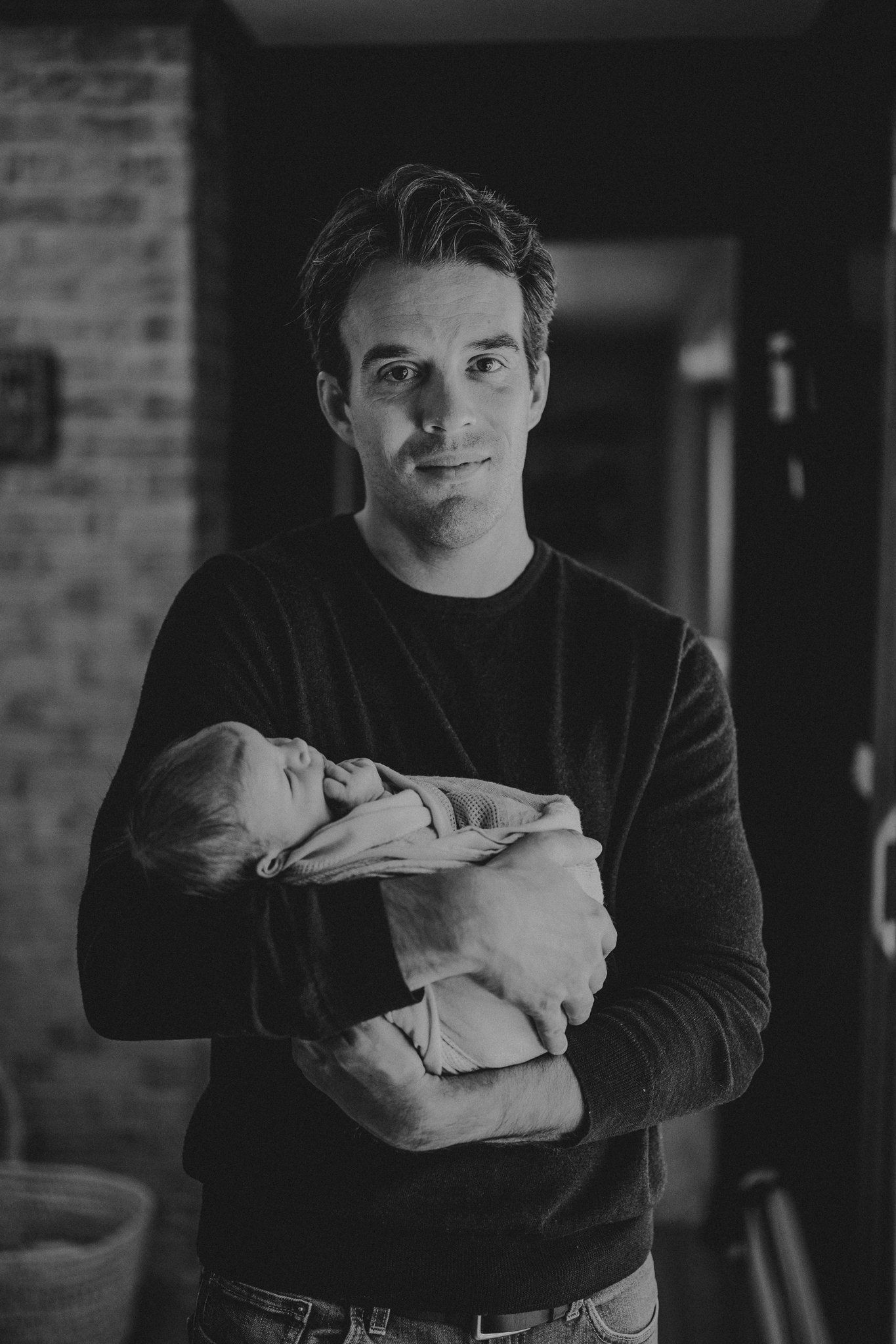 Black and white portrait against brick wall while holding sleeping newborn.