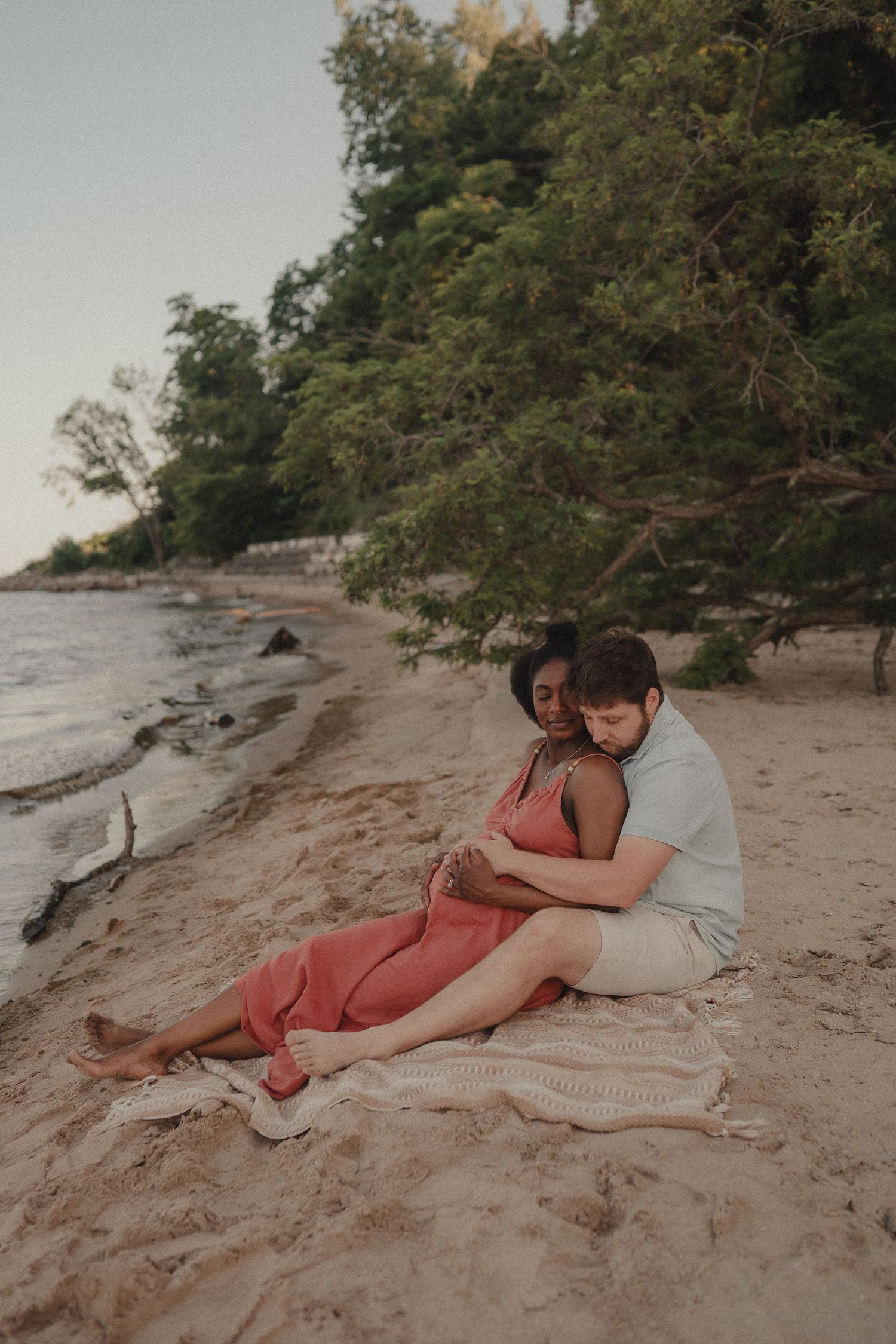 Interracial couple sitting on blanket on sandy beach, man embracing pregnant woman from behind.