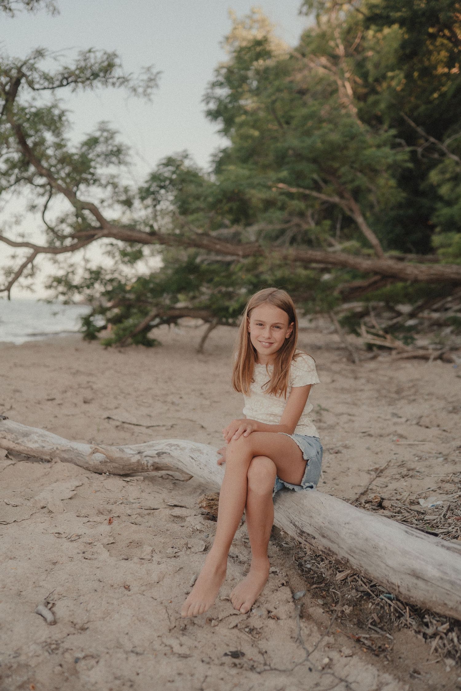 A barefoot girl sits smiling on a driftwood log on a sandy beach surrounded by lush green trees.