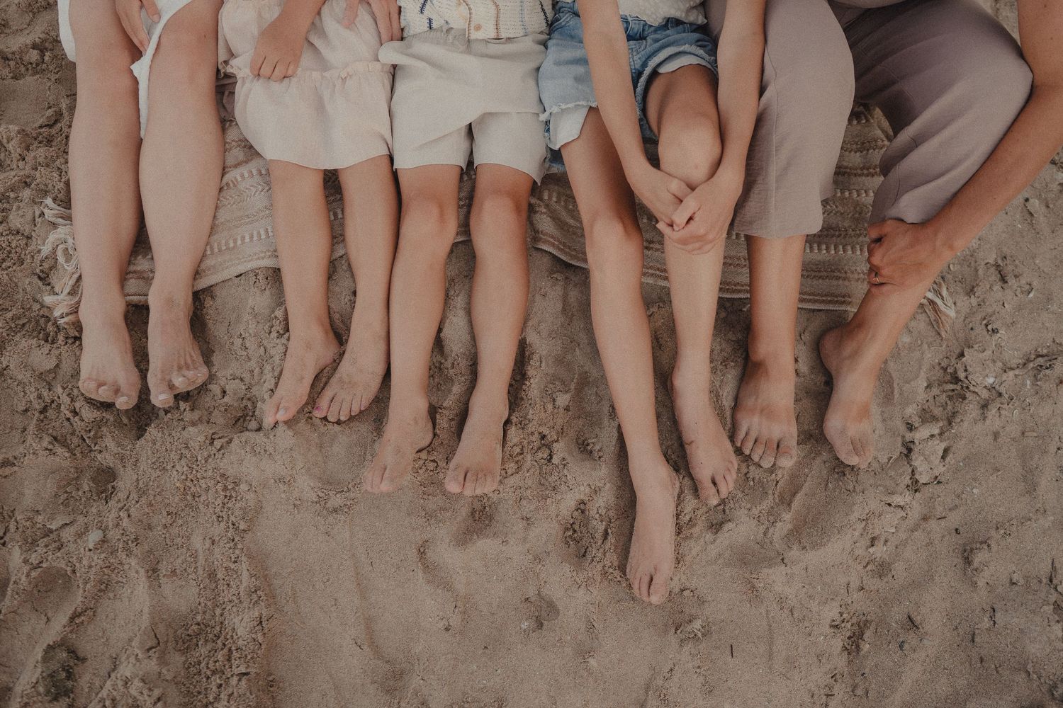 Close-up of five pairs of bare feet resting on sandy beach together.