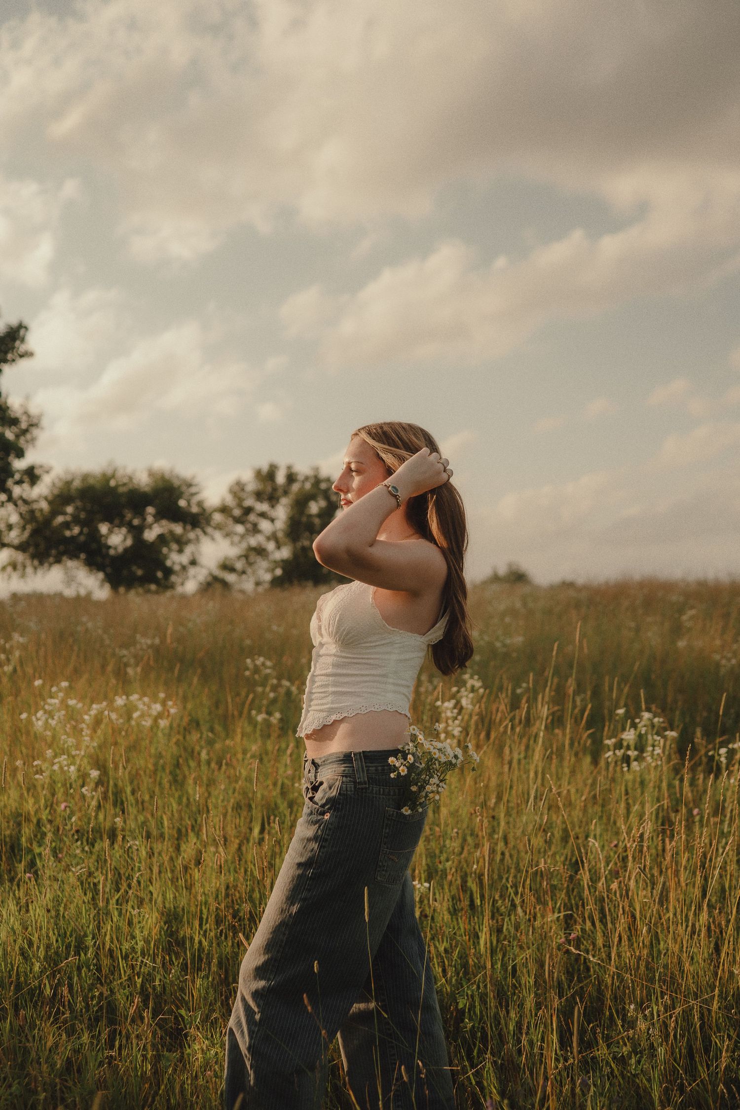 Young woman in white crop top and jeans standing in a wildflower meadow, hand in hair, under a cloudy sky.
