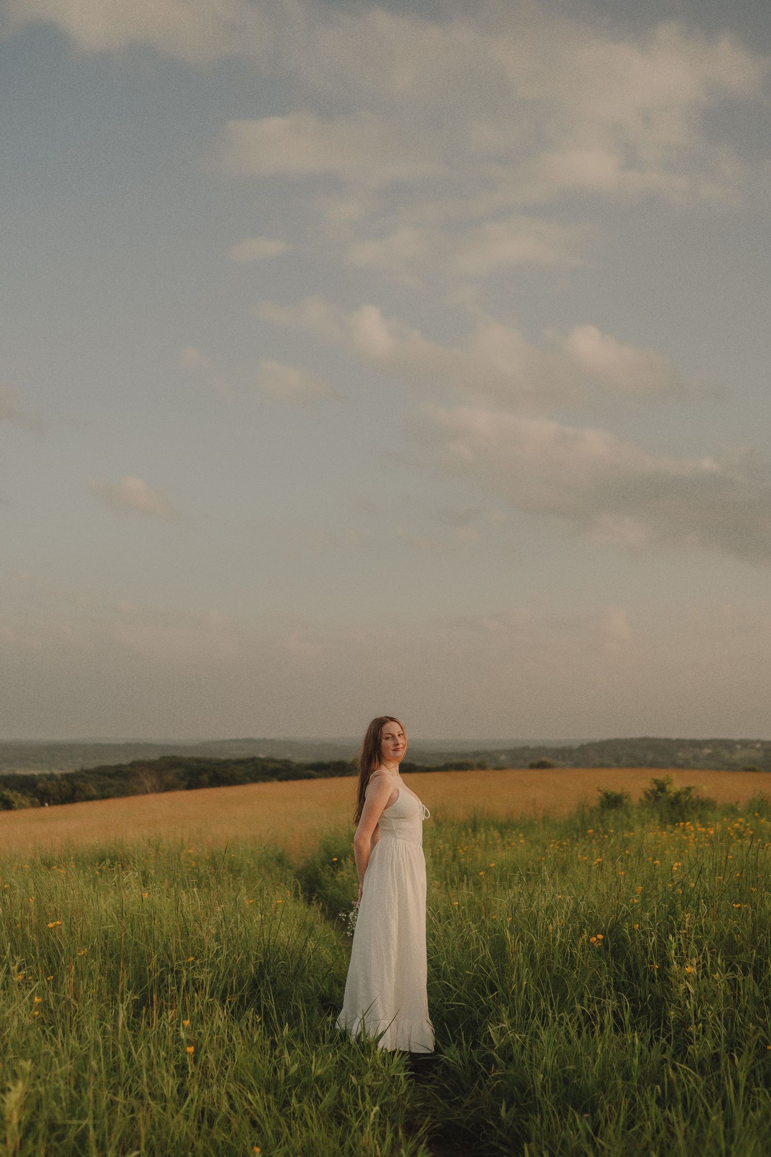 Young woman in a flowing white dress standing in a golden meadow under a dramatic cloudy sky at sunset.