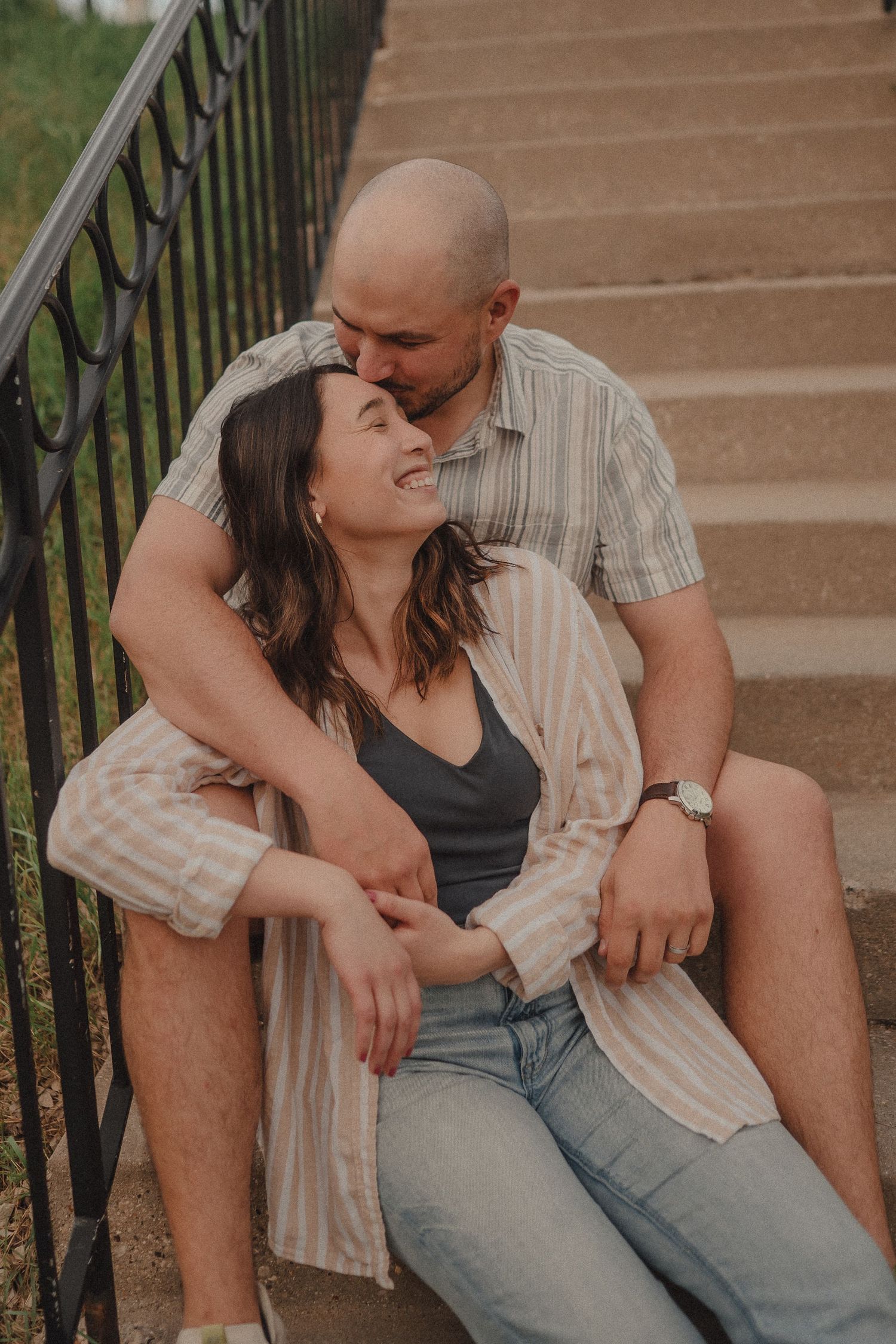 Affectionate couple sitting on outdoor steps, woman laughing as man nuzzles her head.