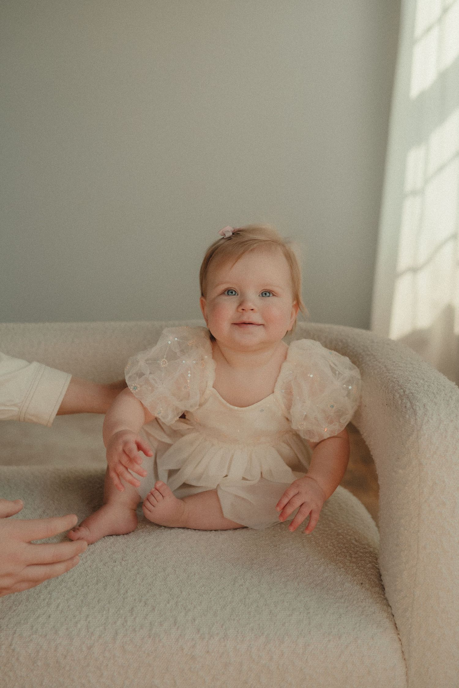 An adorable blue-eyed baby girl in a cream tutu dress sits smiling on a plush chair.