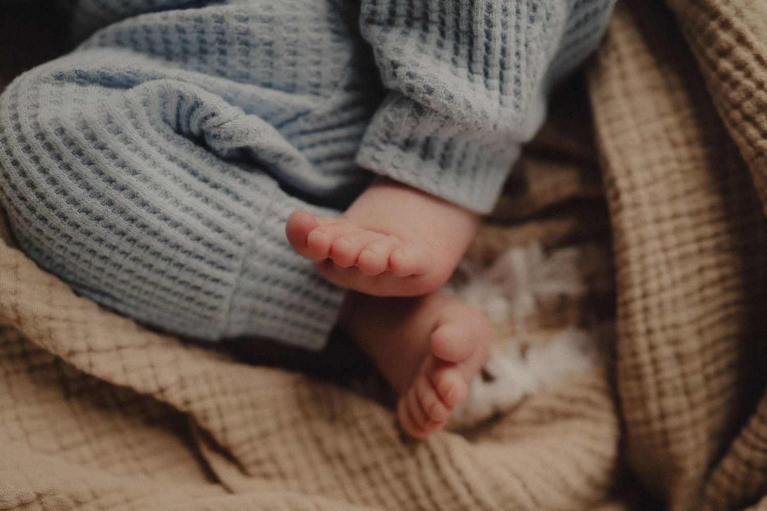 Close-up of newborn baby feet in soft blue knit pajamas resting on a cozy beige blanket.
