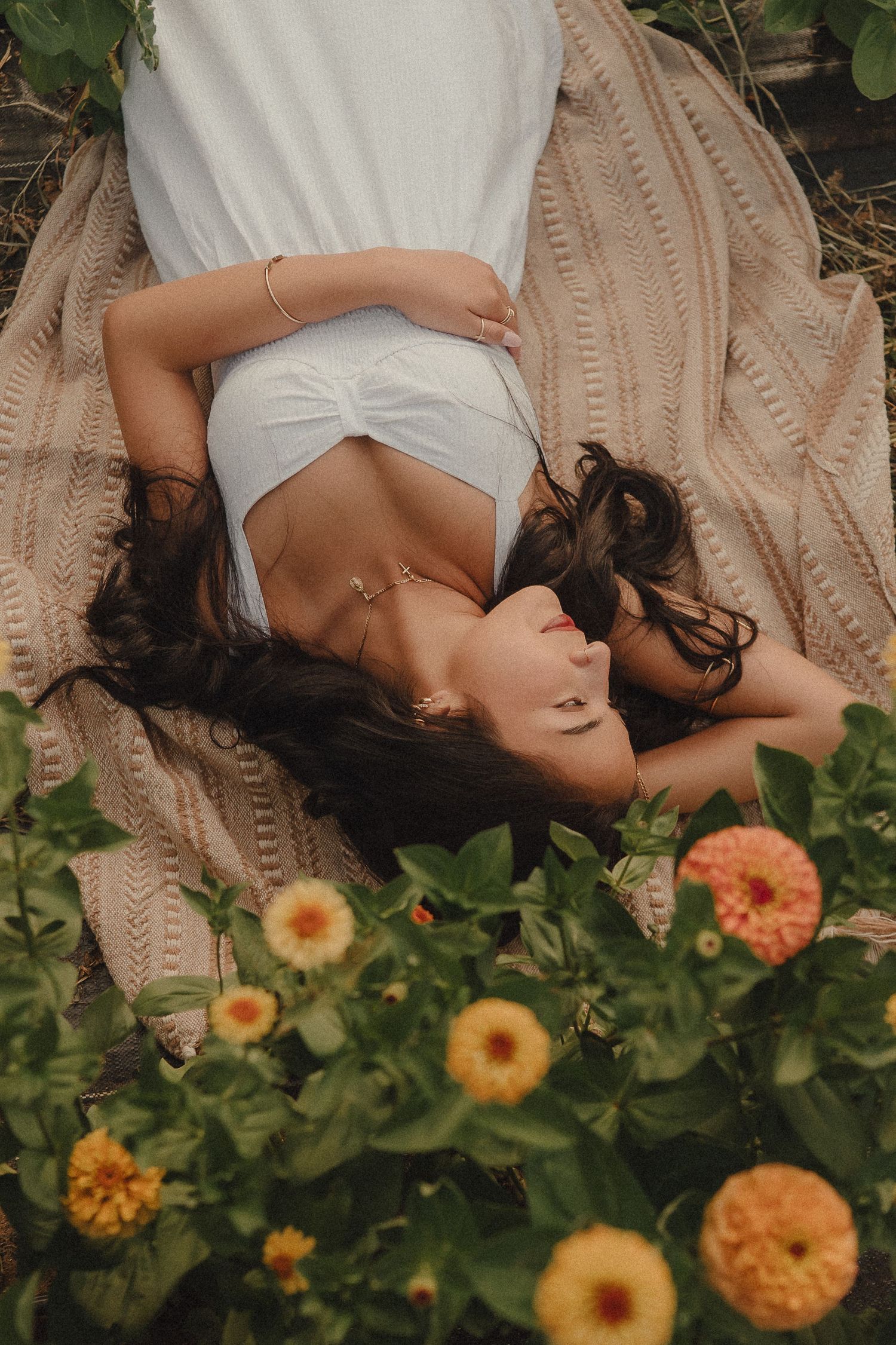 Woman in white dress lying on a blanket surrounded by yellow and orange zinnia flowers in a garden.