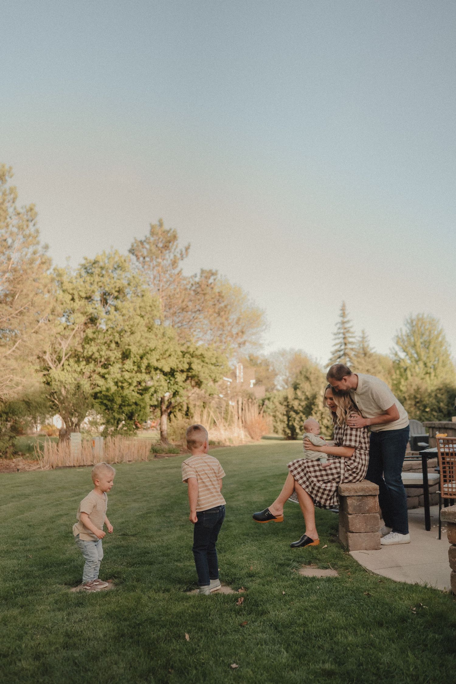 Family of four enjoying golden hour outdoors, parents sharing a moment while two boys play on grass.