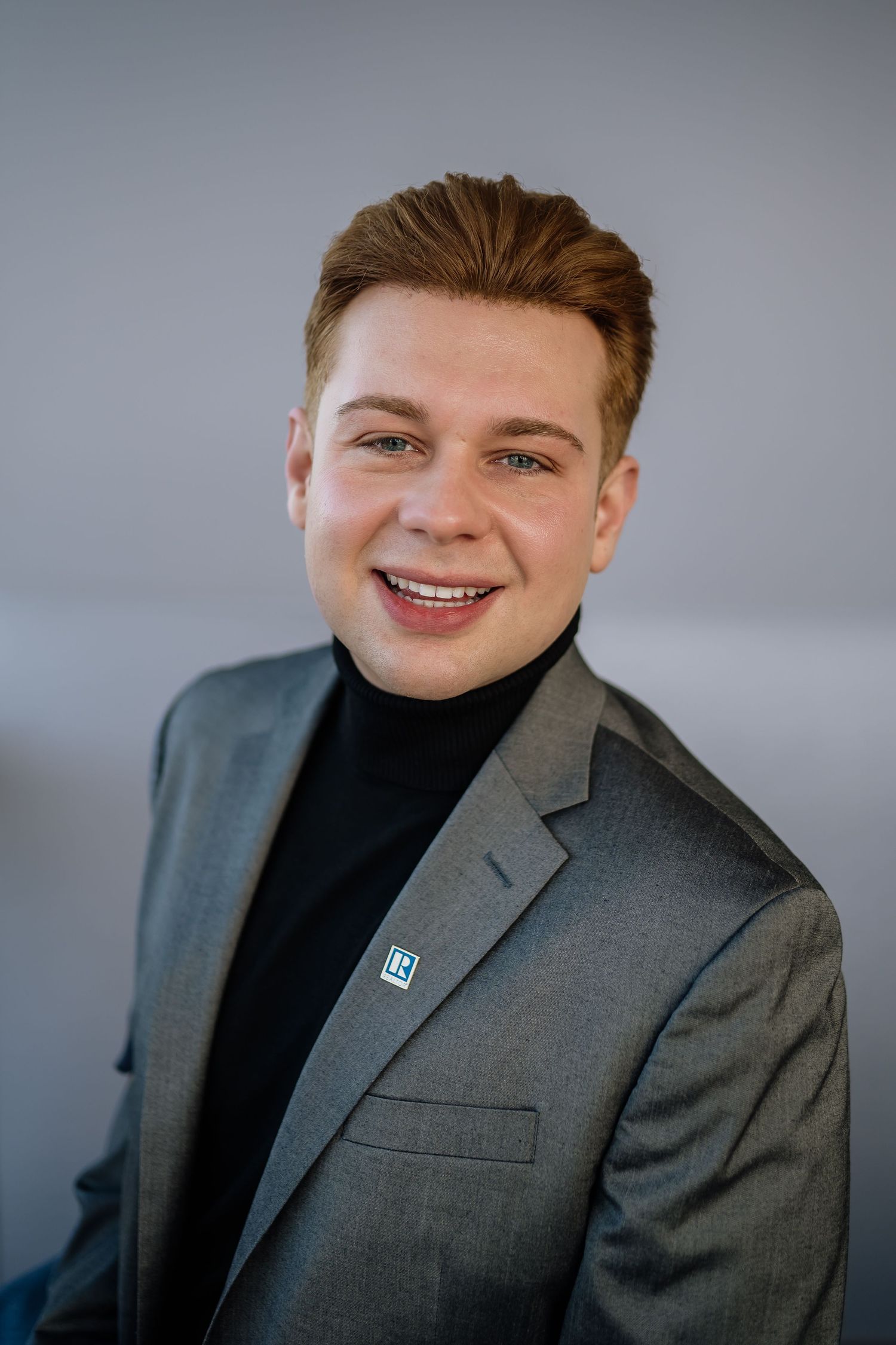 Professional portrait in grey blazer and black turtleneck against blue background.