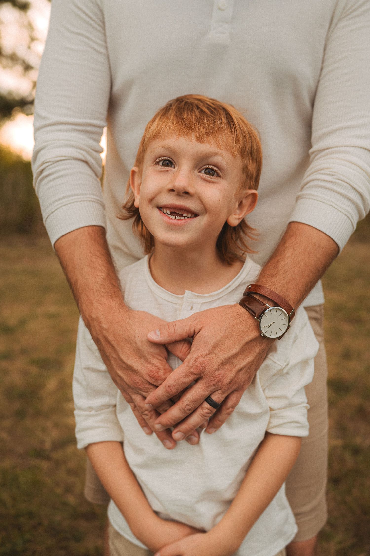 Child wearing white shirt with adult's hands on shoulders at sunset.