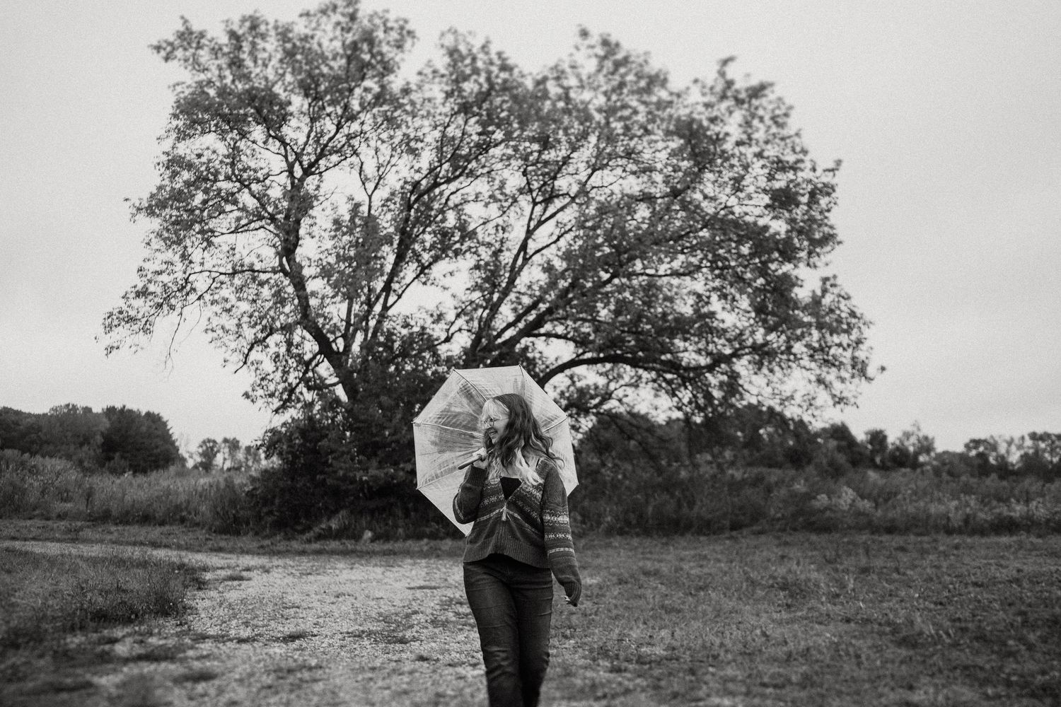 A black and white shot of someone twirling with an umbrella in an open field with a large tree.