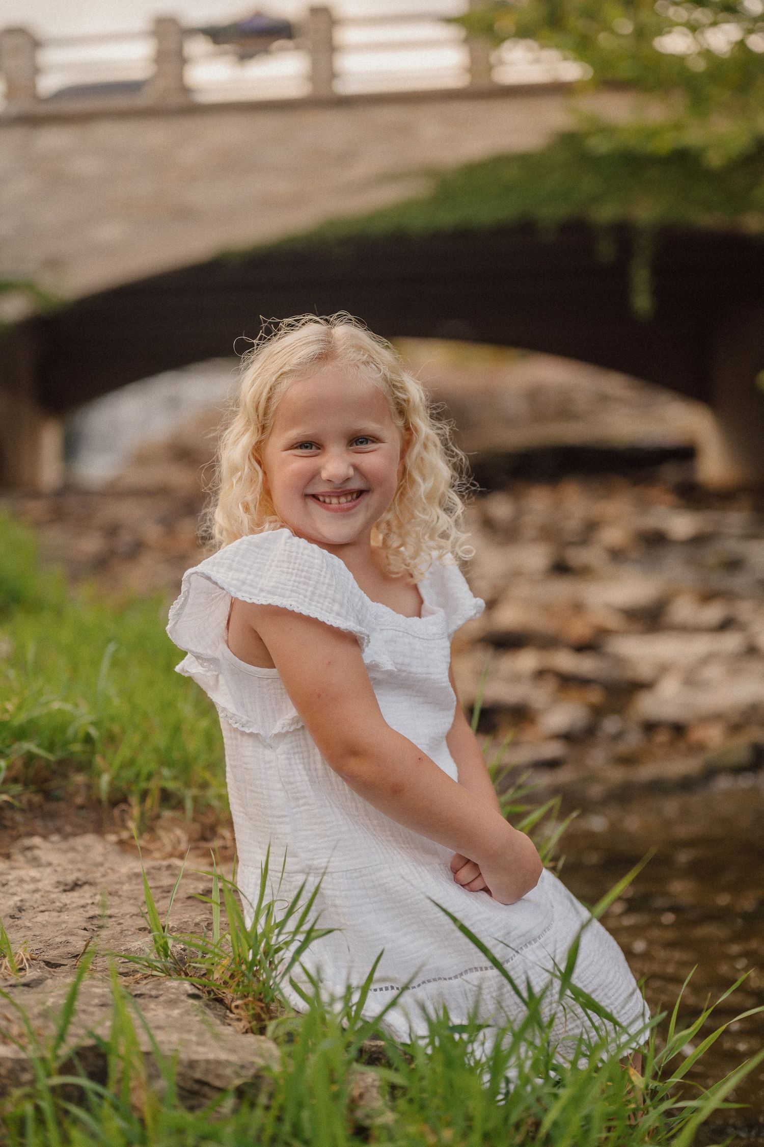 Young girl in white dress plays and laughs near a stone bridge and stream.