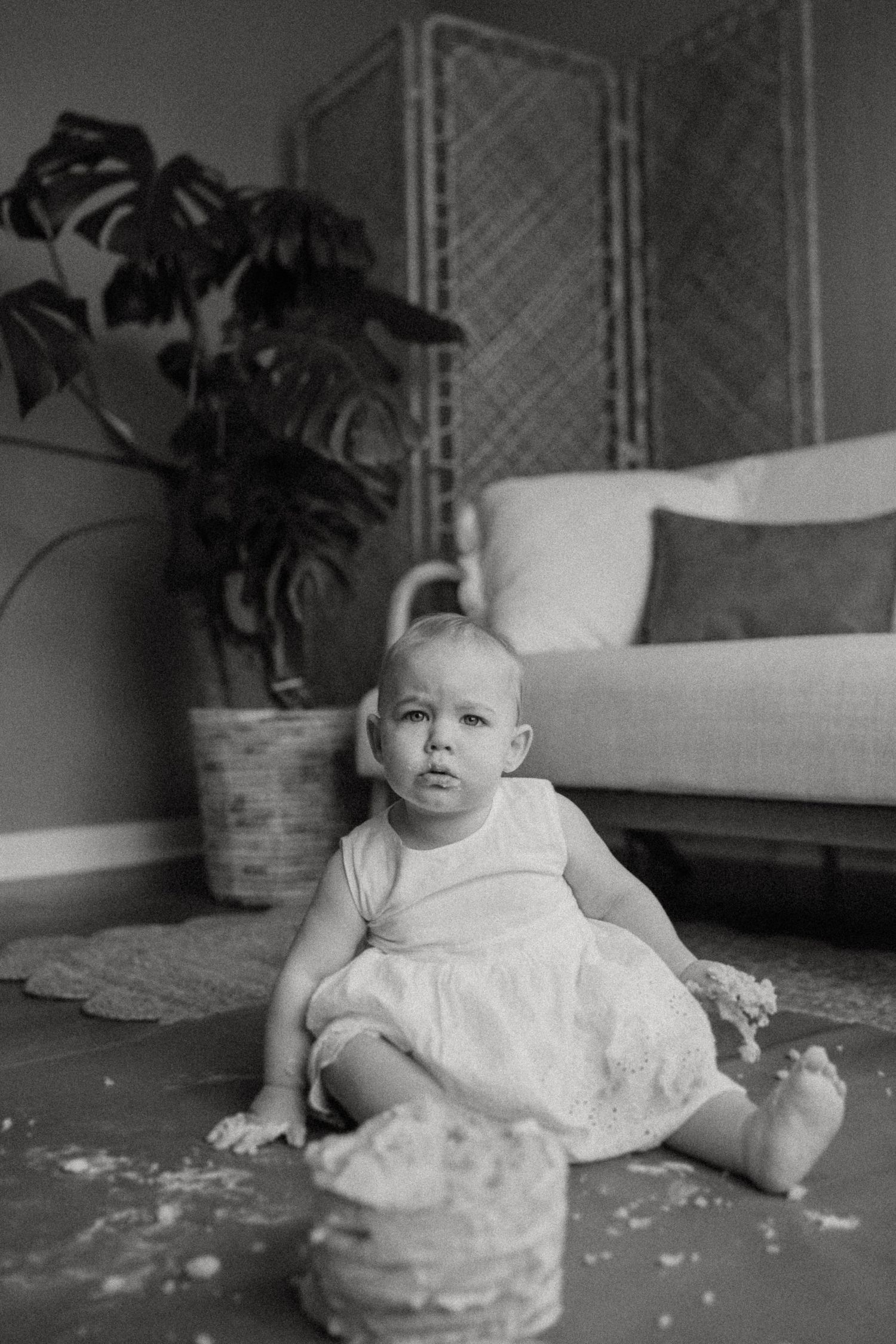 A toddler in white sitting on the floor with a smashed cake and houseplant backdrop.