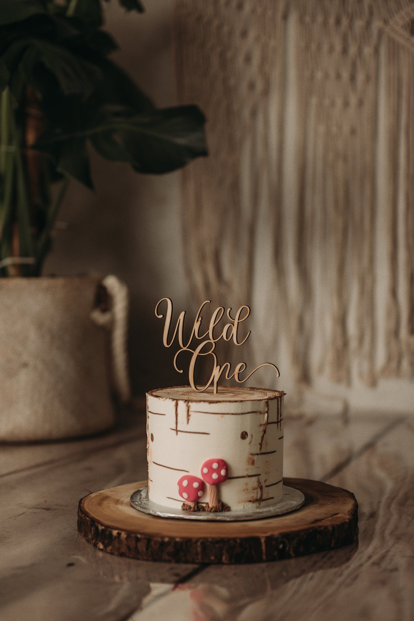 A rustic woodland cake with 'Wild One' topper and red mushroom decorations sits on a wooden slice platter.