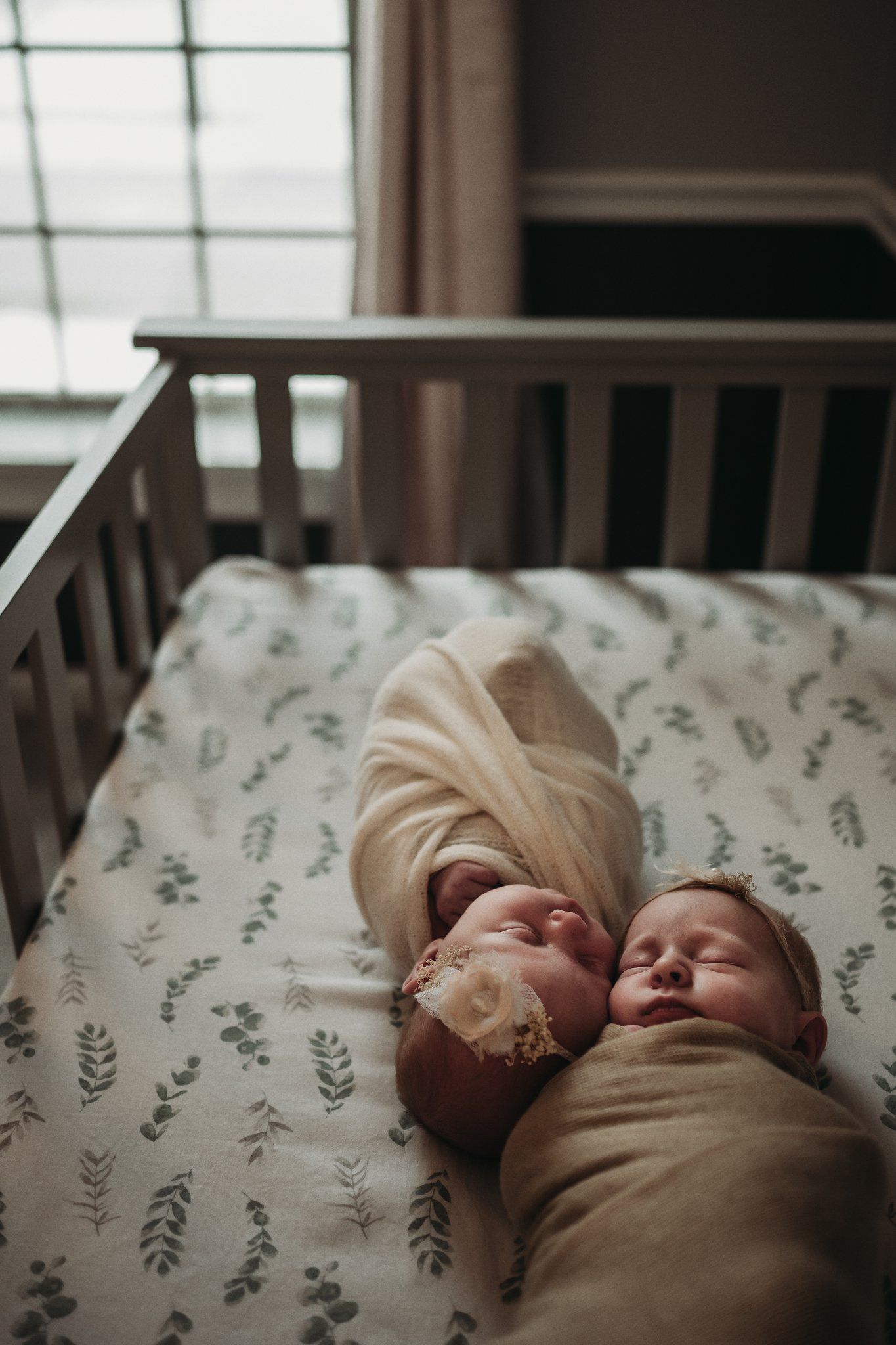 Two swaddled babies sleeping side by side in wooden crib with floral bedding.