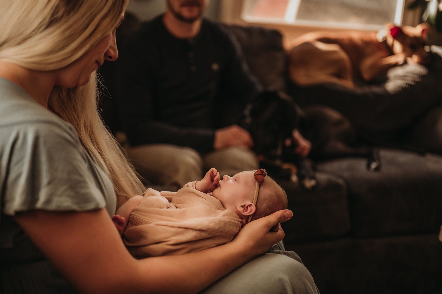 A quiet moment on a dark couch as a sleeping newborn is cradled in soft lighting.