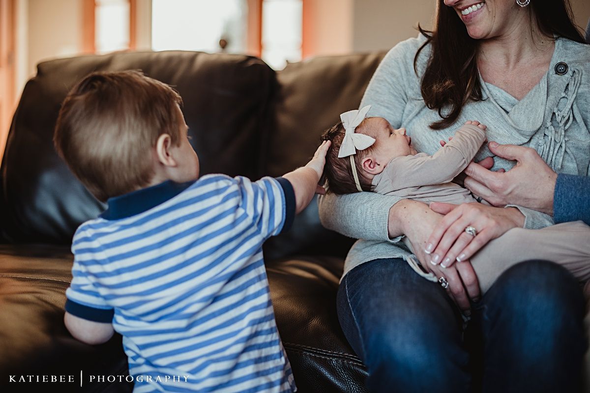 A toddler in striped shirt reaches out while sitting on a brown leather couch during an indoor family photoshoot.
