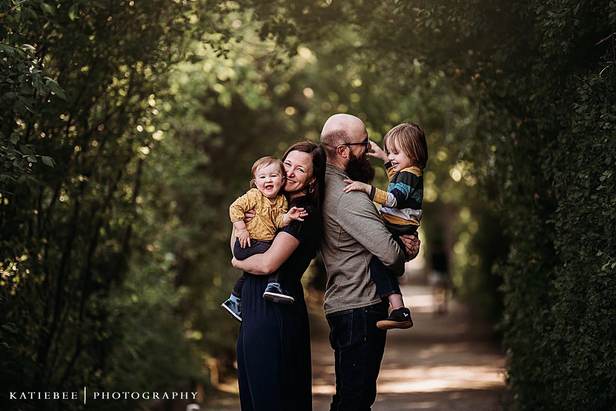 Family portrait in a sunlit garden path surrounded by lush greenery and bokeh lighting effects.