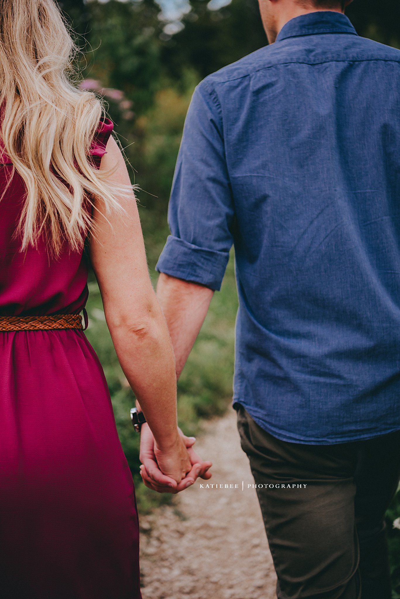Close-up of couple holding hands, with burgundy dress and blue shirt against nature backdrop.