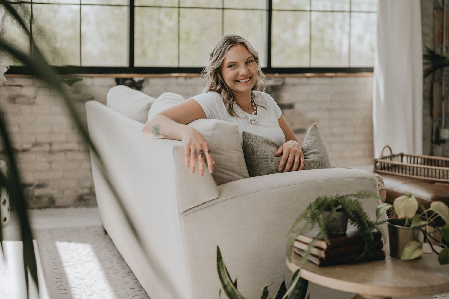Relaxed pose on white armchair in sunny room with industrial windows and plants.