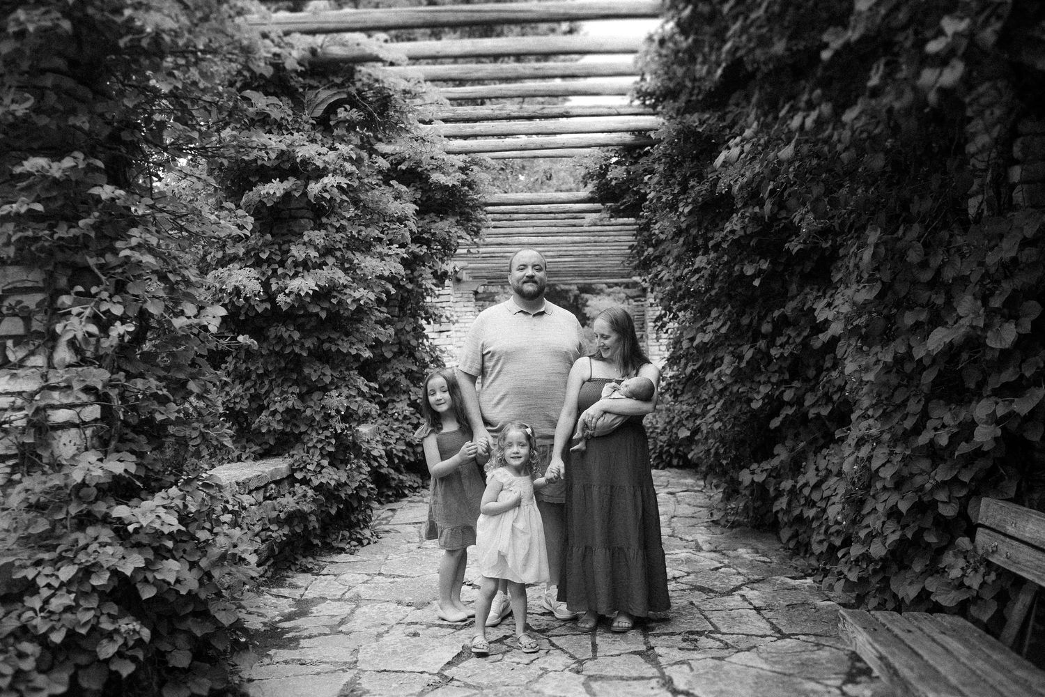 A family stands together under a garden pergola surrounded by lush hedges in black and white.
