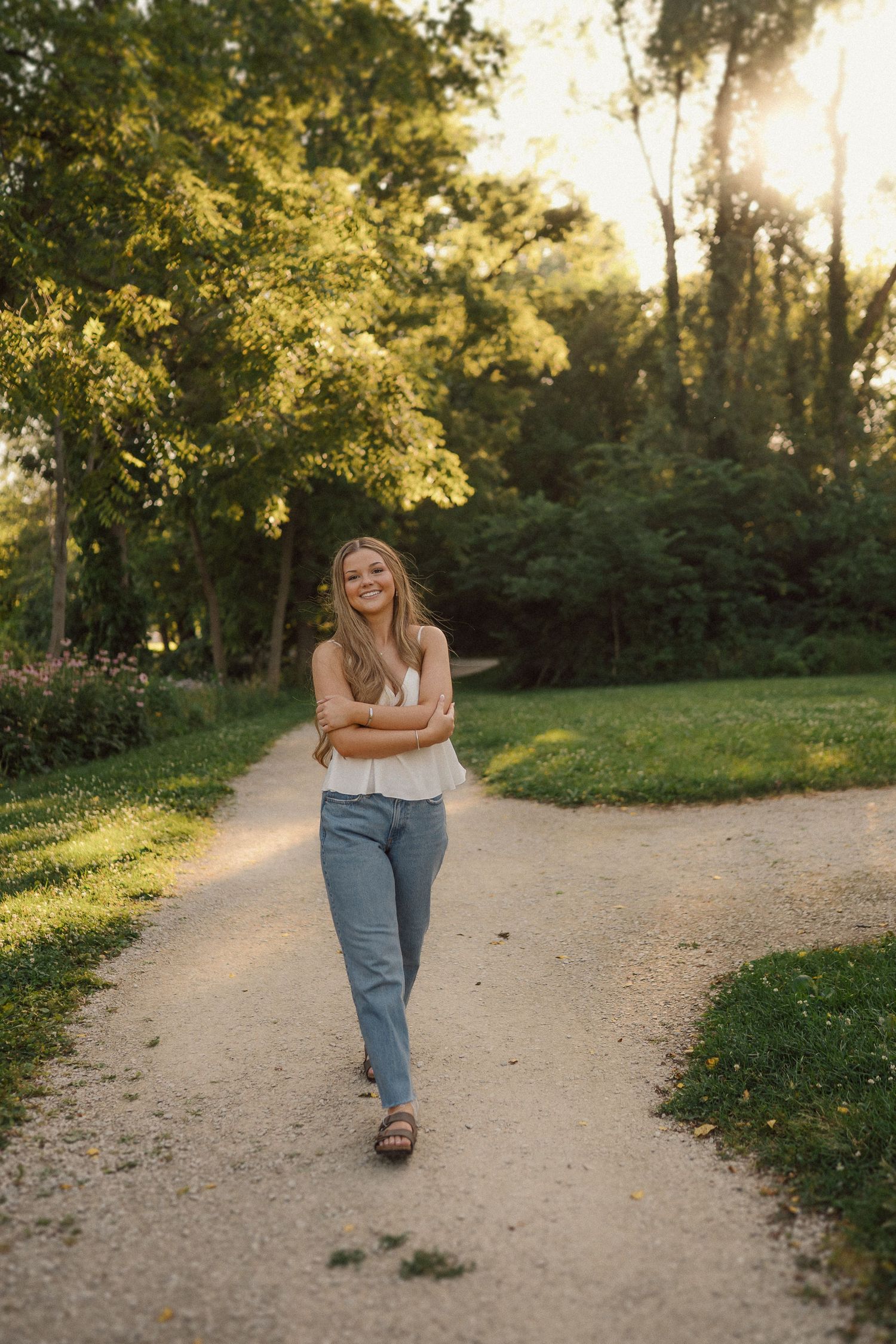 A woman strolls down a sunlit path wearing jeans and a casual top in a serene park setting.