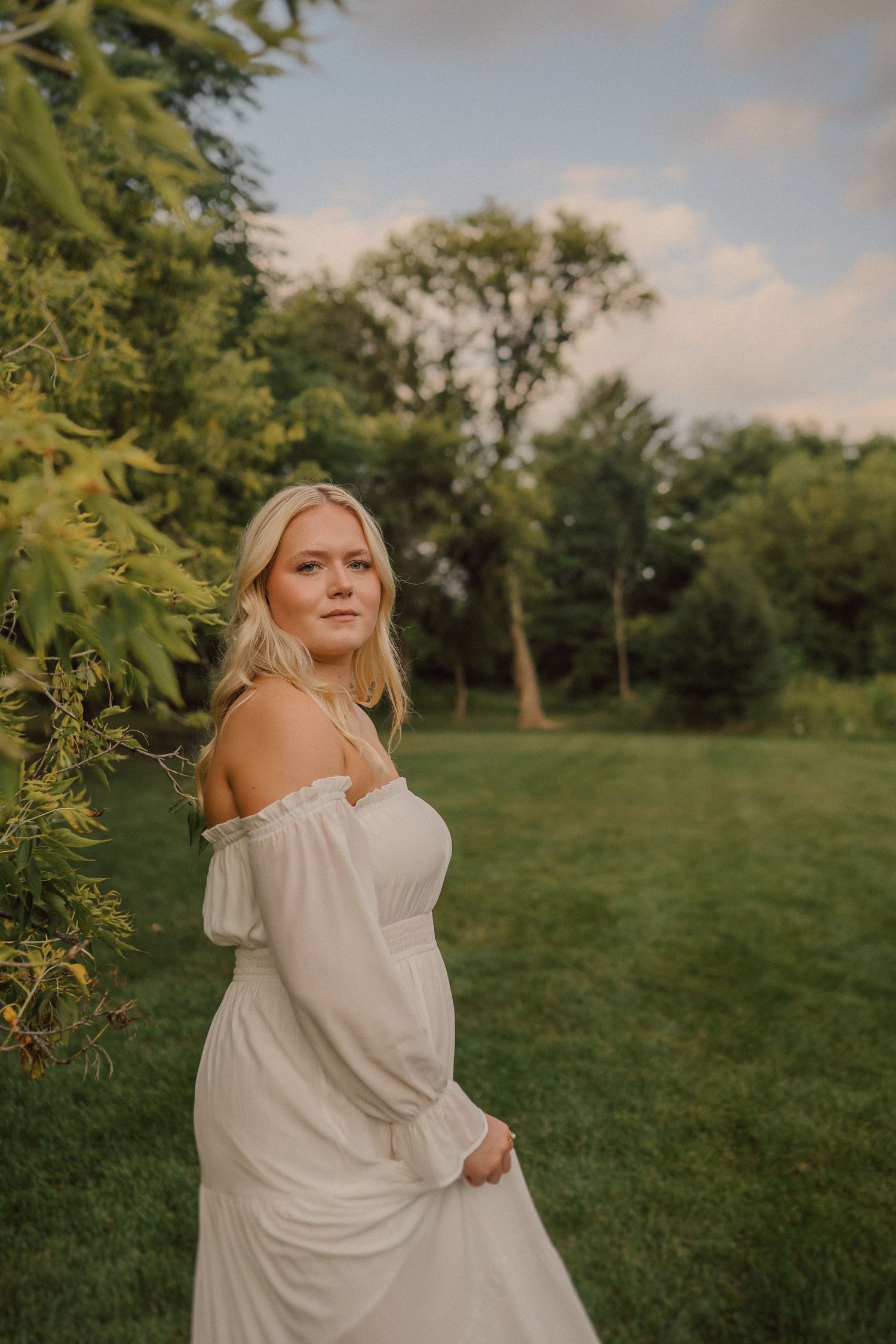 Person in white off-shoulder dress poses gracefully in a sunlit garden setting.