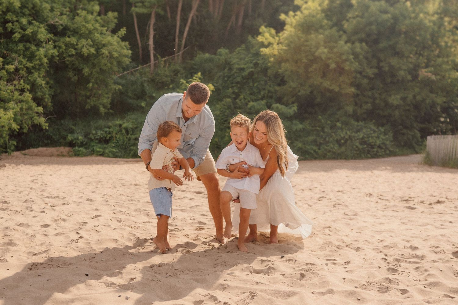 Family wearing white and beige clothing plays together on sandy beach at sunset.