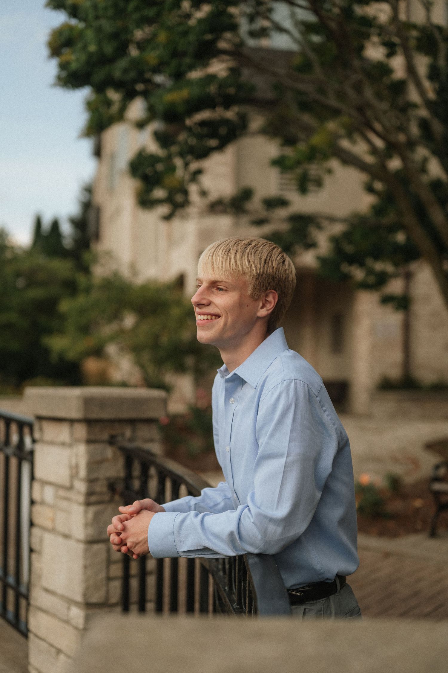 A person in a light blue dress shirt smiles while standing near stone pillars and railings outdoors.