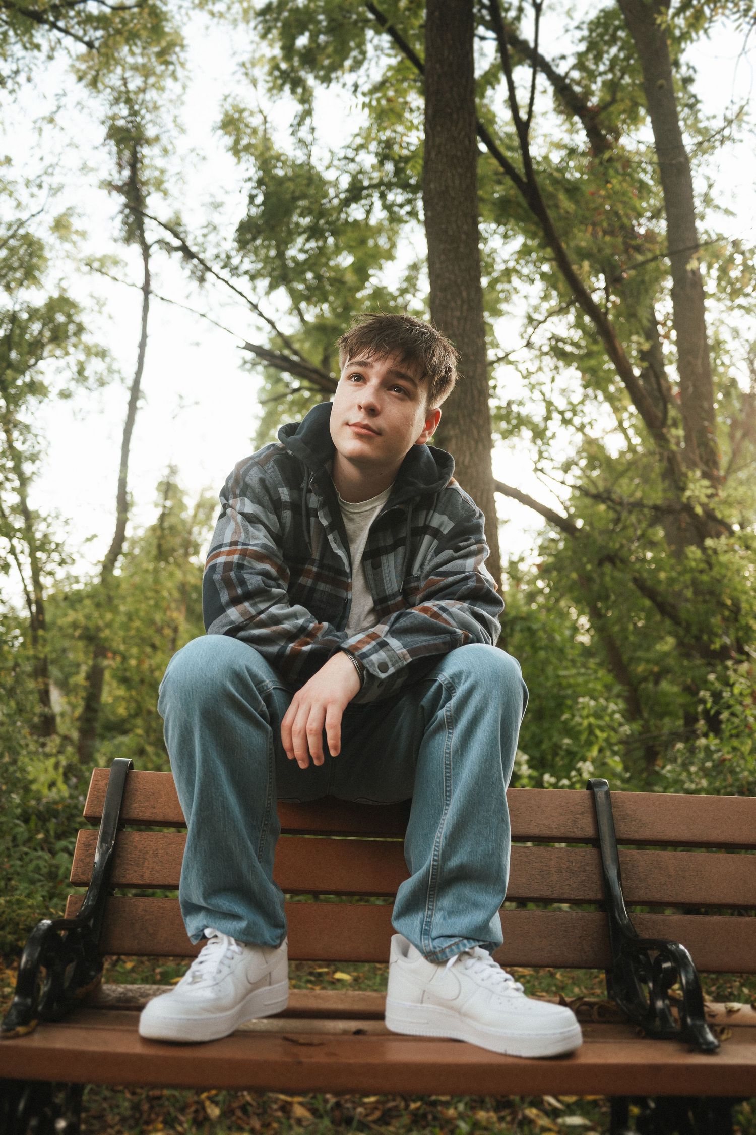 A casual portrait of someone sitting on a wooden bench in a park wearing a plaid jacket and jeans with white sneakers.
