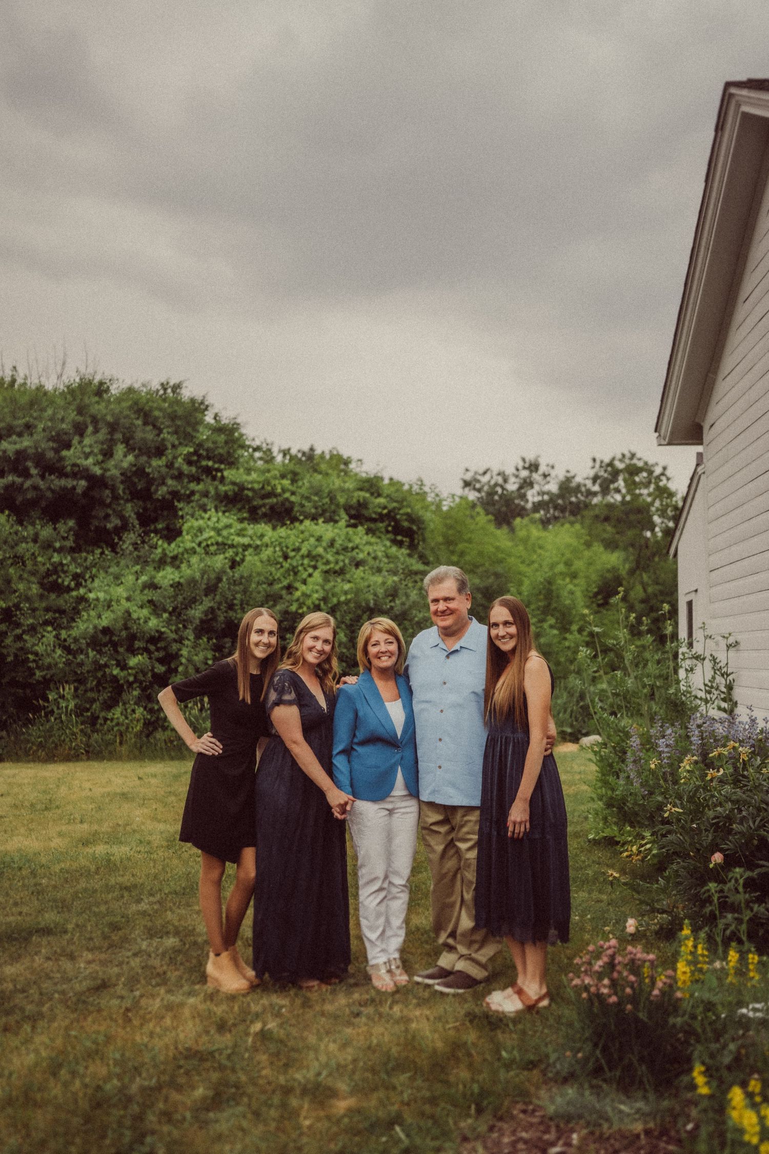 Group photo outside a white farmhouse with flowering gardens and green trees in background.