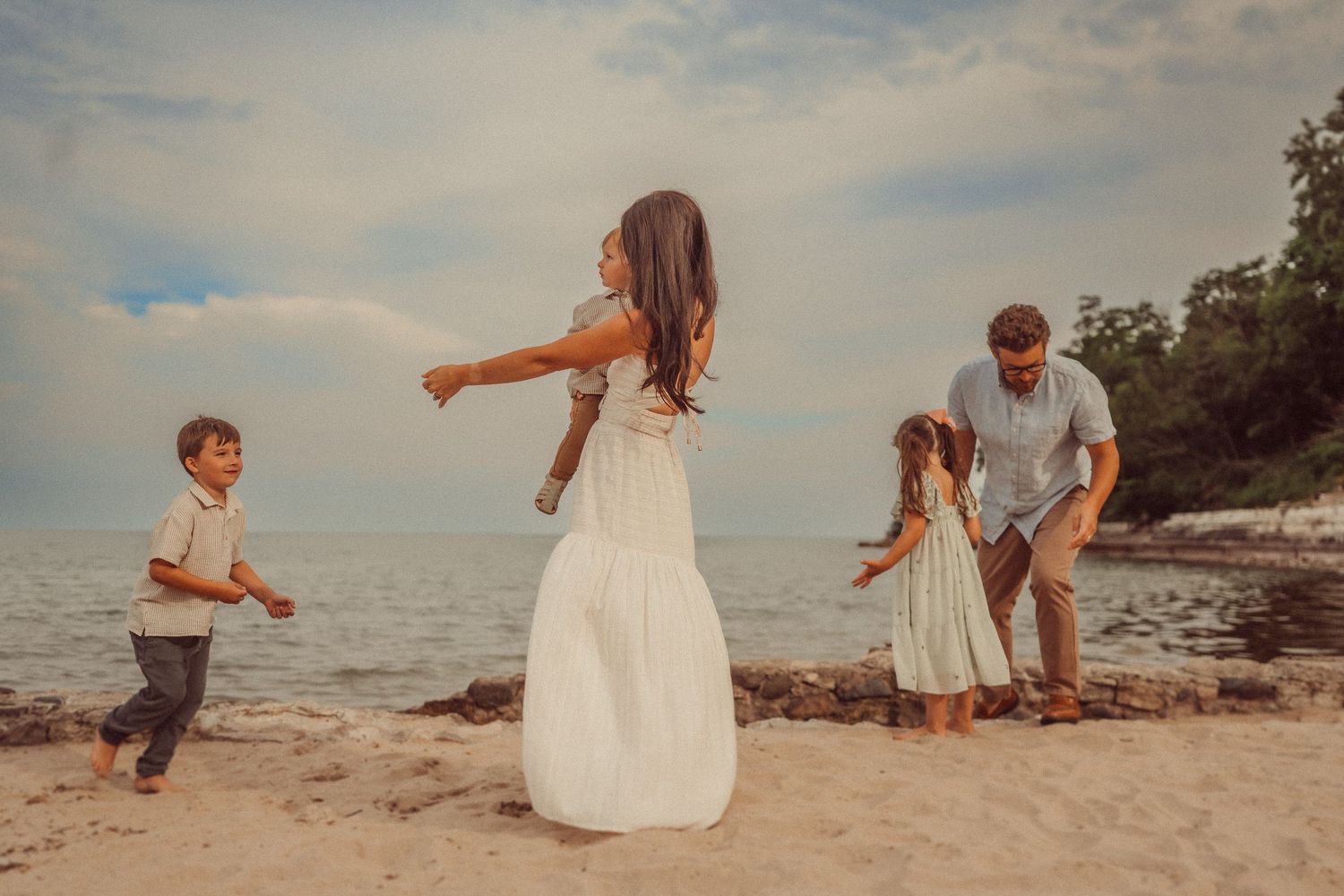 A family plays together on a sandy beach at sunset near rocky shores and calm waters.