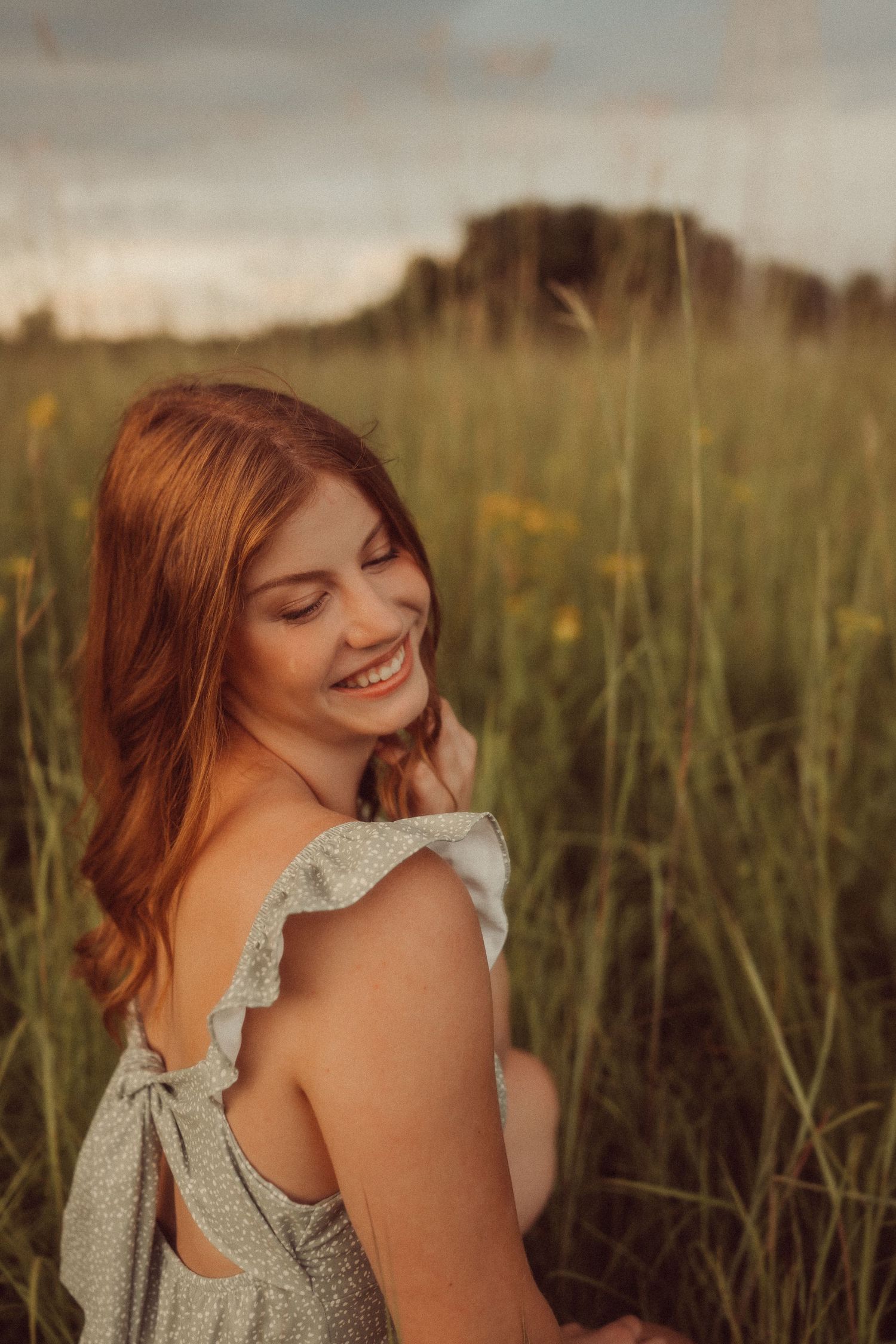 Woman in a floral dress smiling in a sunlit meadow with tall grass and wildflowers during golden hour.