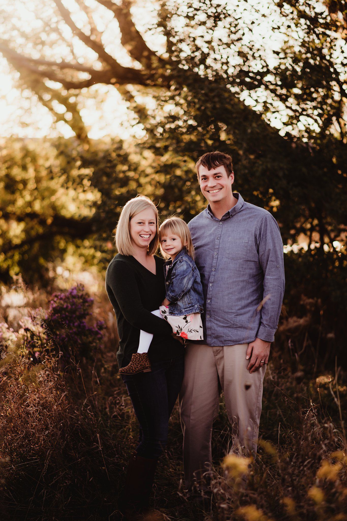 A family portrait captured in warm autumn light with trees and golden foliage in the background.