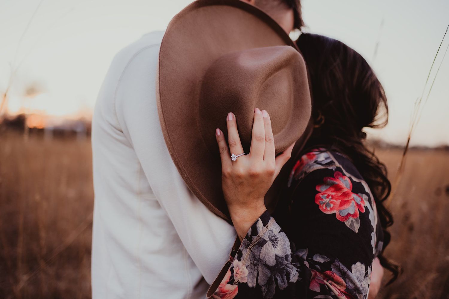A brown felt hat covers two people sharing an intimate moment in a rustic field setting.