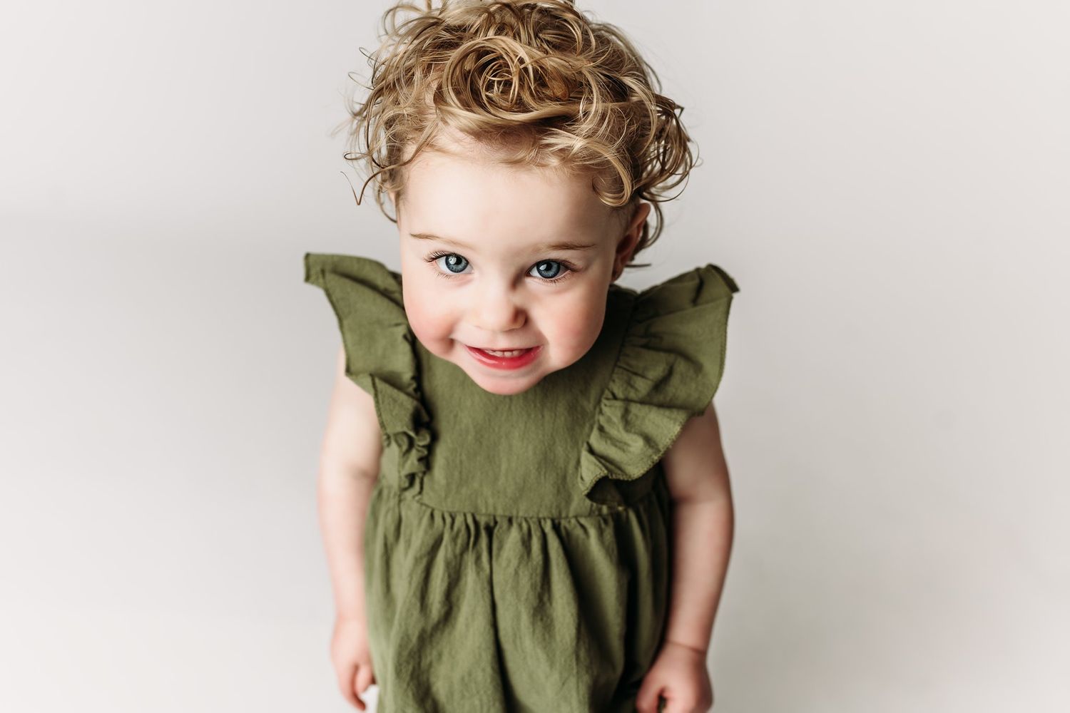 Child in olive dress smiles sweetly at camera against light backdrop.
