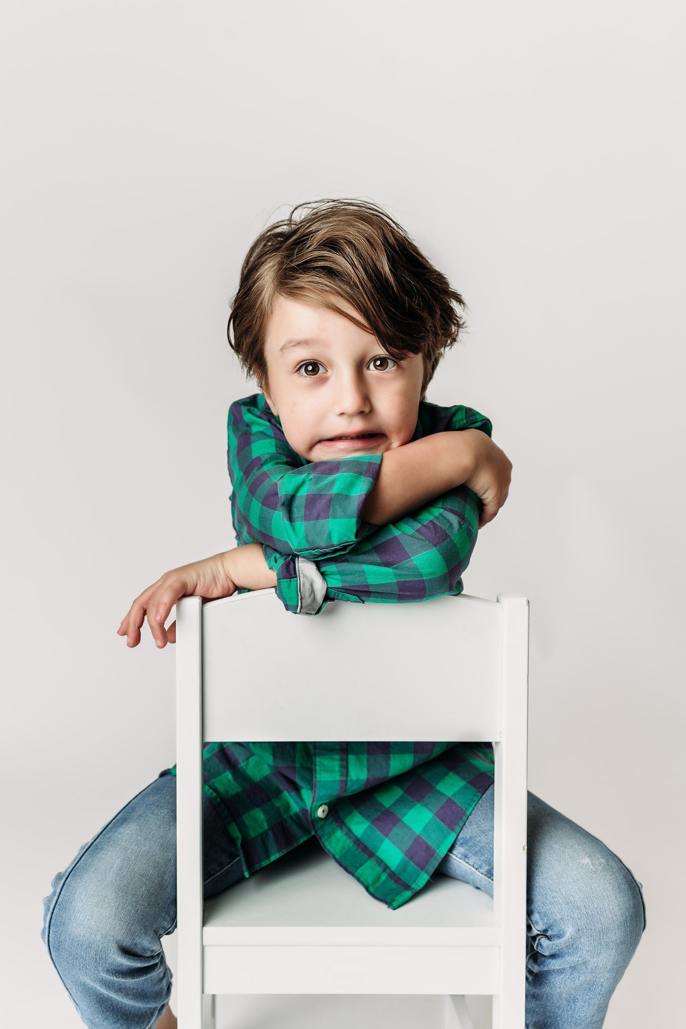 Child in green checkered shirt rests arms on white chair back with contemplative expression.