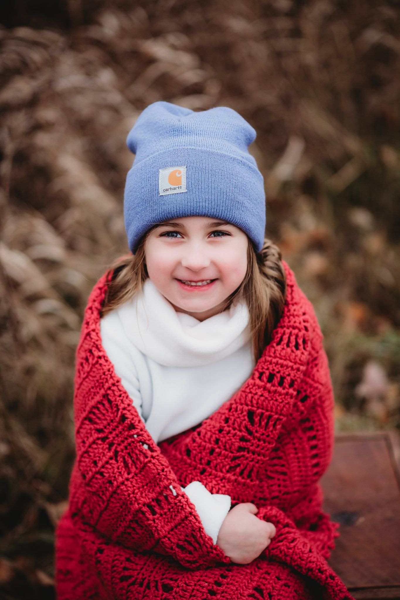 A child wrapped in a red knit blanket and blue beanie smiles warmly at the camera.