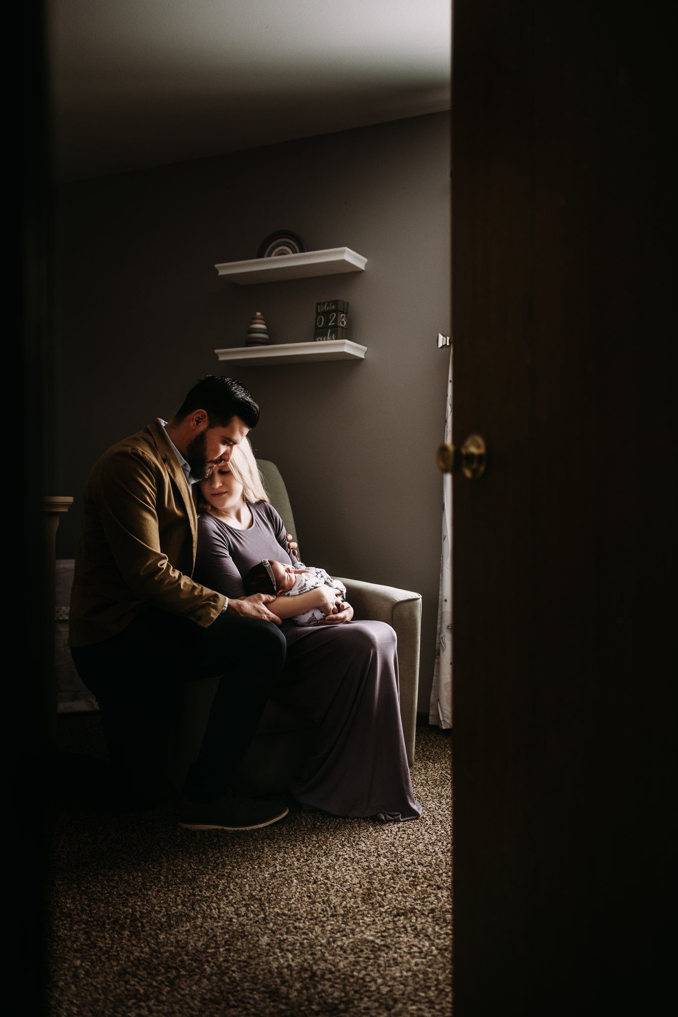 Moody dramatic portrait of someone holding a baby in a dimly lit room with floating shelves on the wall.