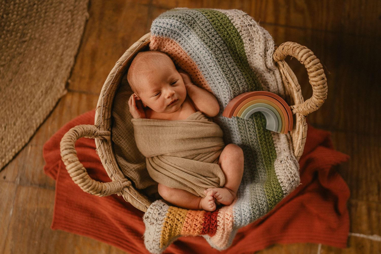 Newborn baby swaddled in beige fabric sleeping peacefully in a basket with knitted blanket and orange backdrop.