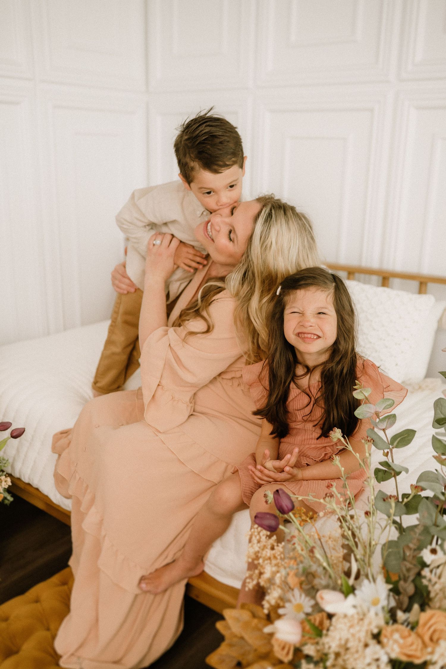 A tender family moment captured on a bed with soft peach tones and delicate floral decor in the foreground.