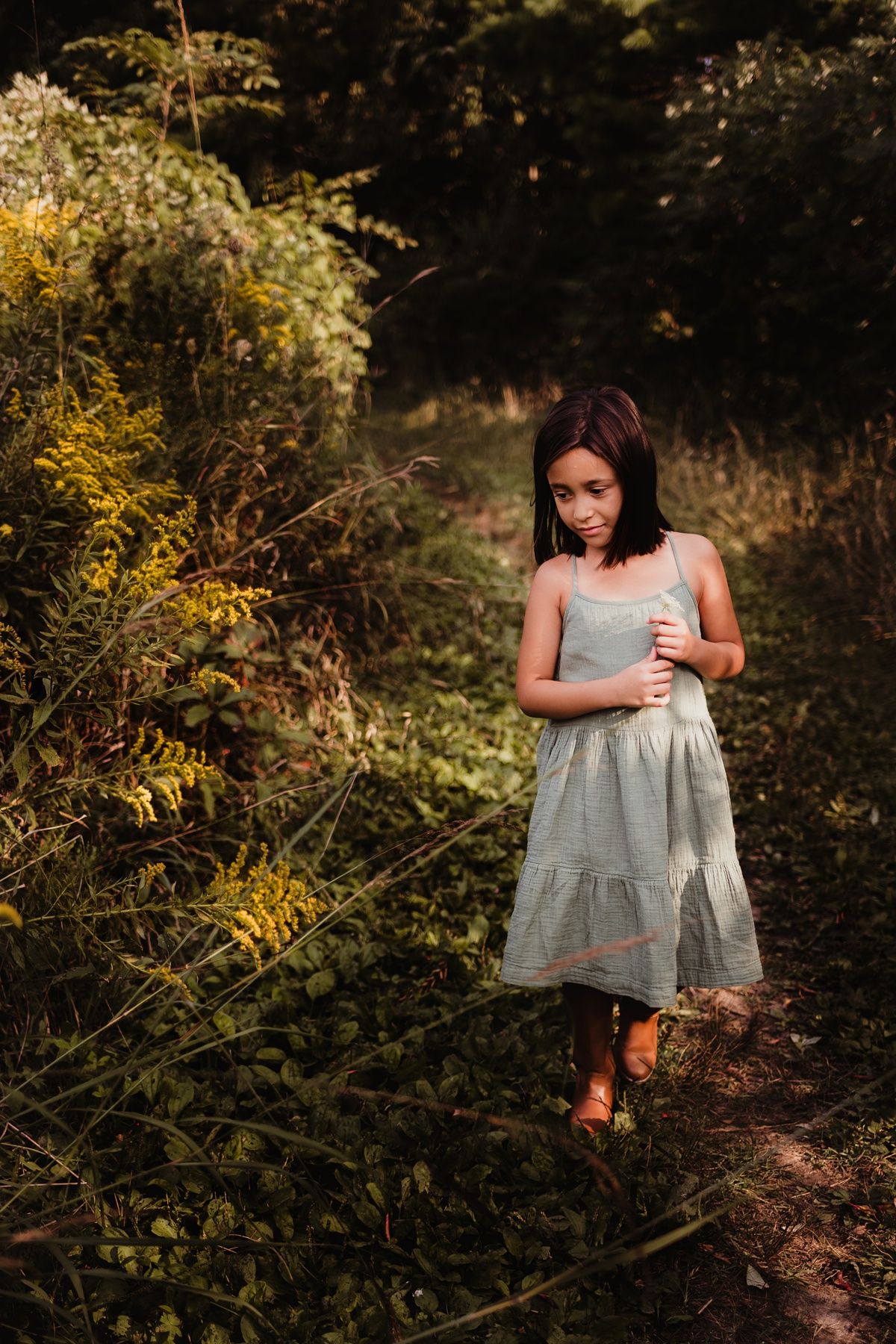 Person in a gray dress walking along a sunlit forest path surrounded by golden foliage.