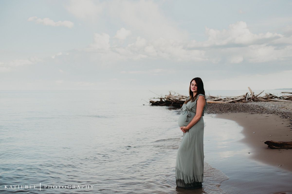 Serene maternity photo of woman in flowing dress standing by ocean at sunset.