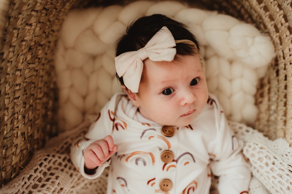 Baby wearing white bow headband and patterned outfit rests in woven basket.
