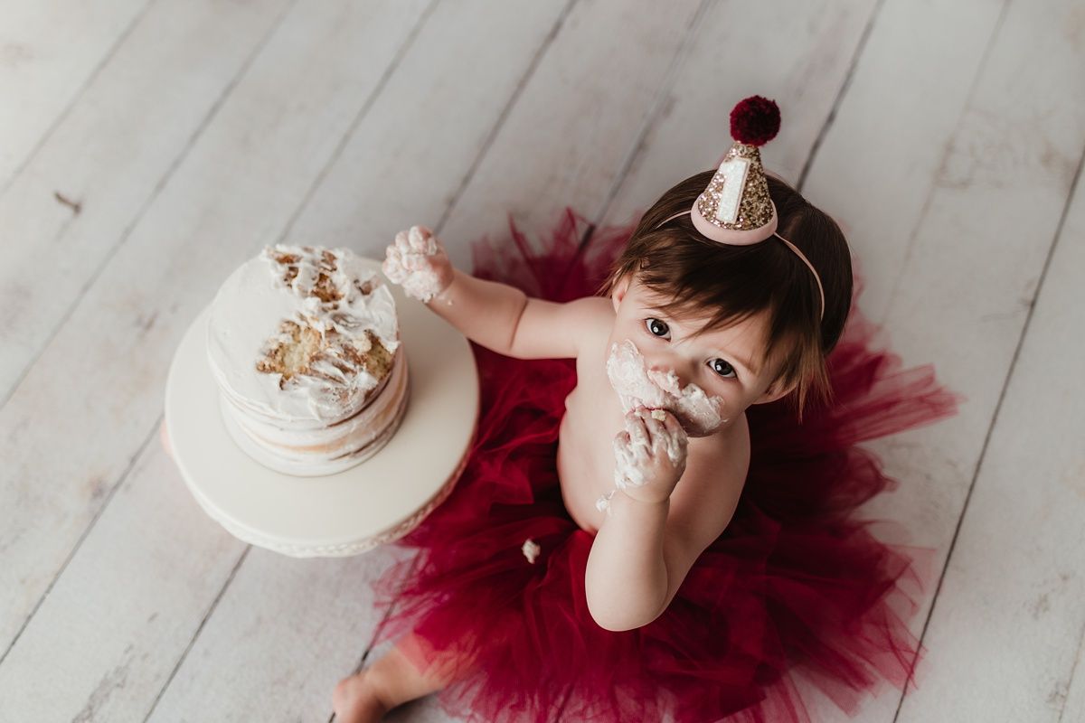 Baby in burgundy tulle skirt and gold party hat plays with birthday cake on wooden floor.