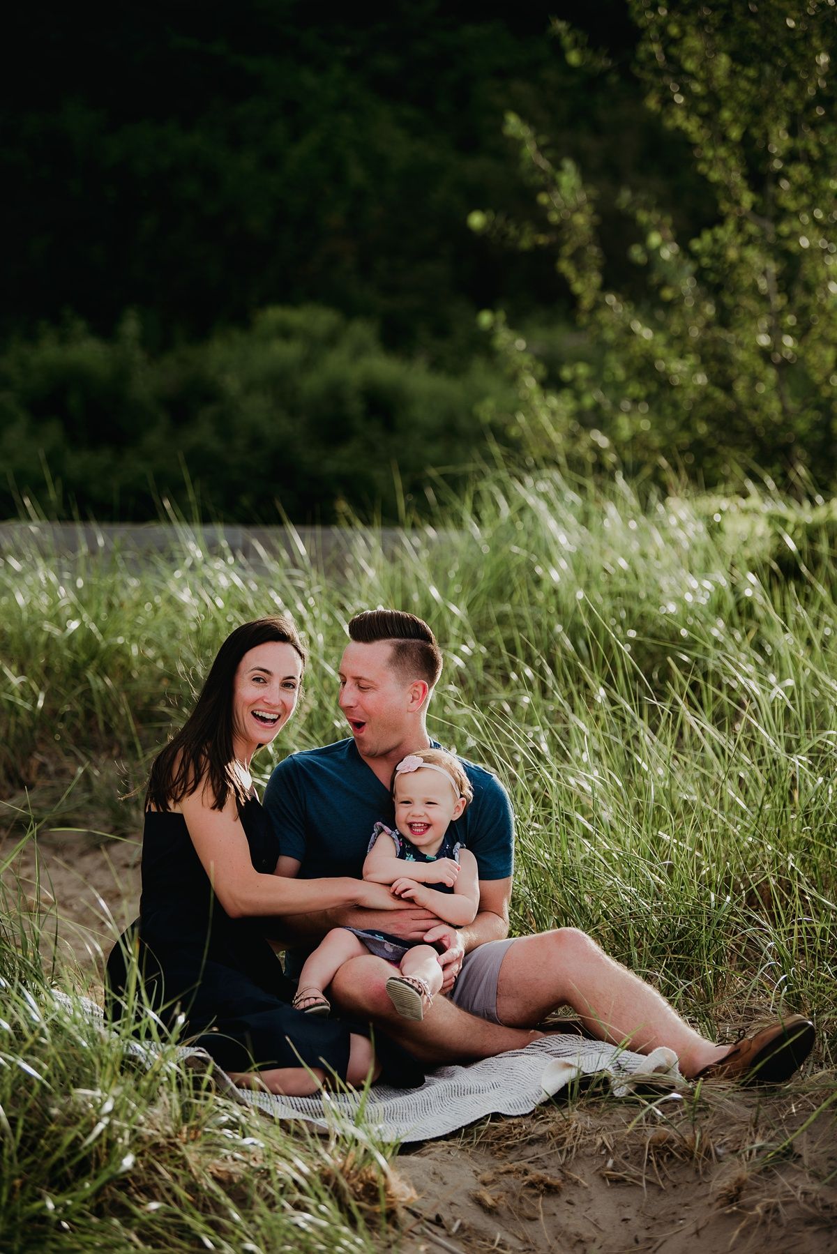 A family sits together on a blanket by tall grass during a summer photoshoot.