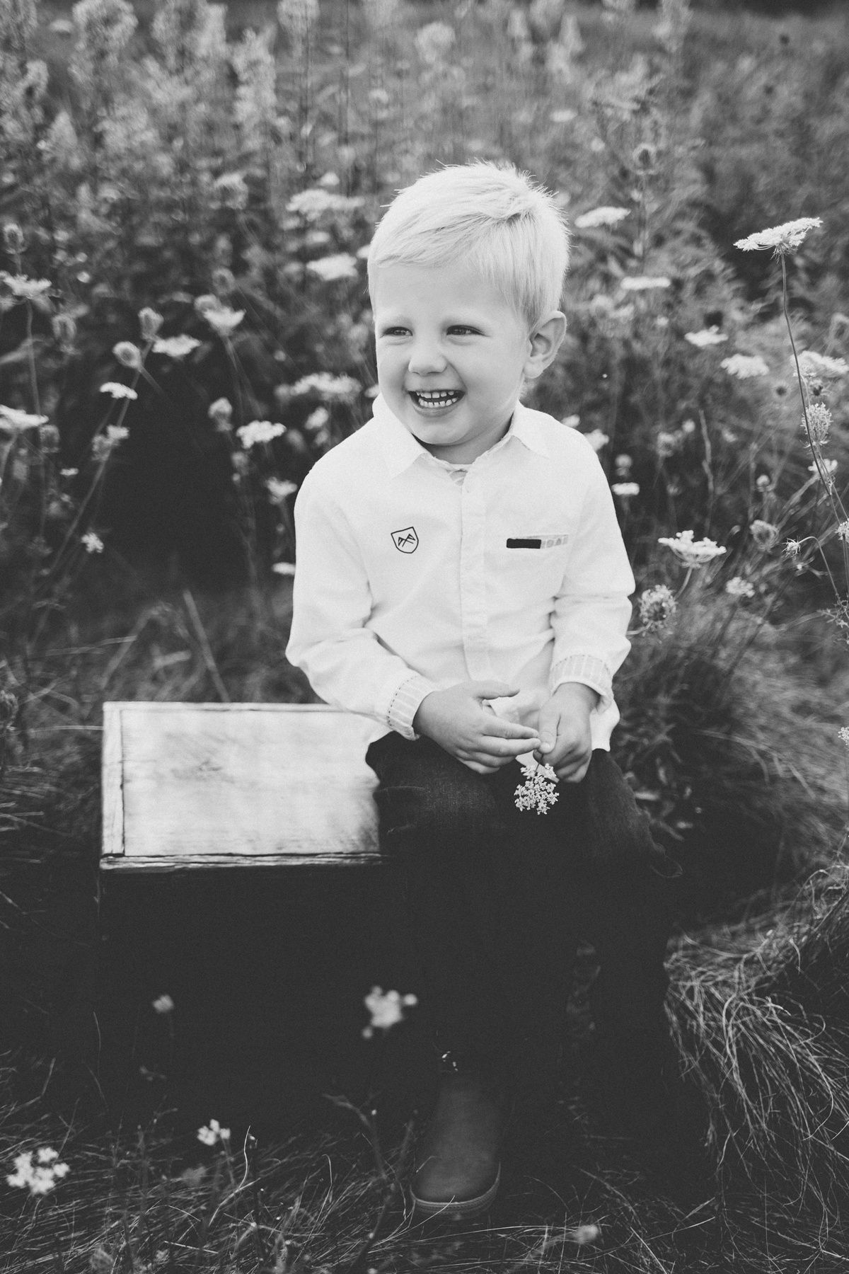 Black and white portrait of child sitting on wooden box in field of wildflowers.