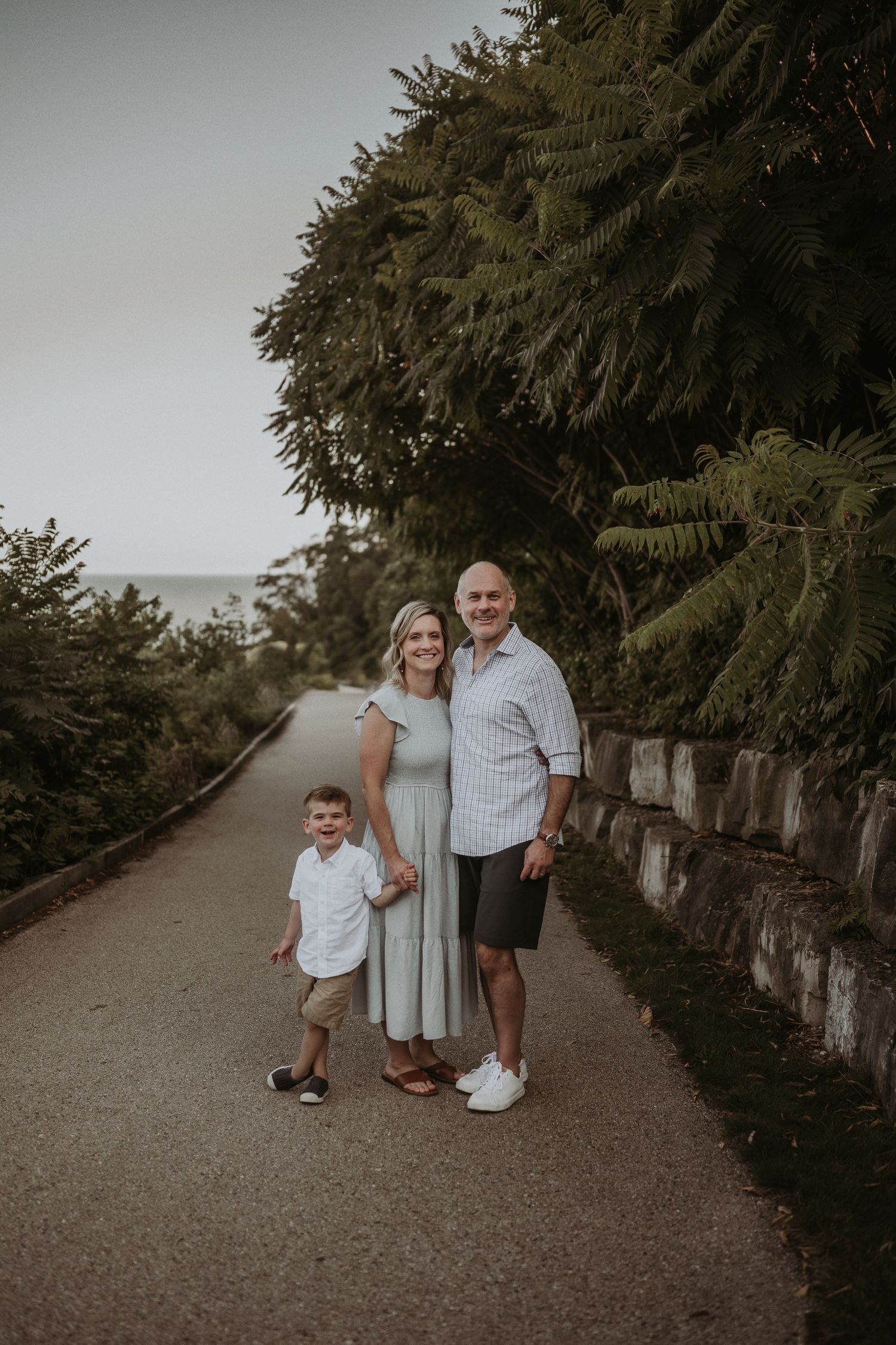 A family stands together on a scenic path with tall evergreen trees and stone wall.