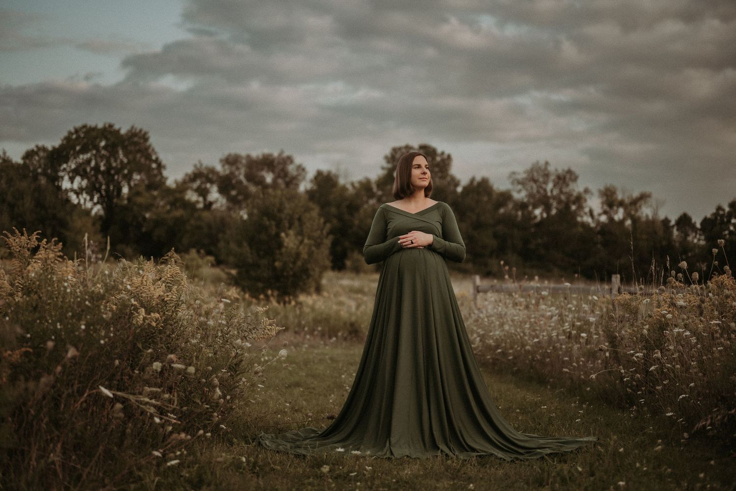 Dramatic maternity portrait of woman in flowing green dress standing in autumn field.