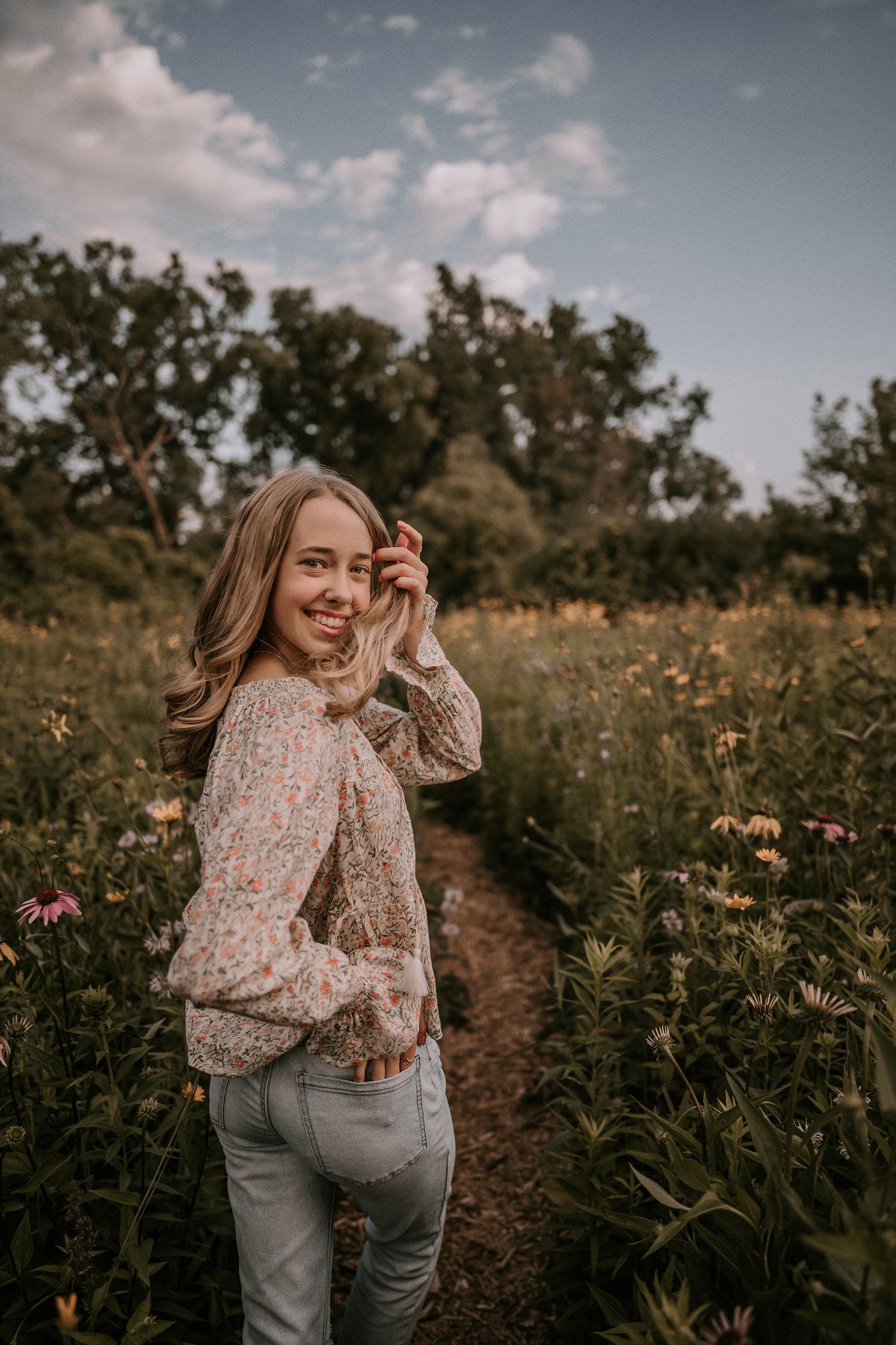 A candid portrait of someone in a floral blouse turning back to smile in a wildflower field.